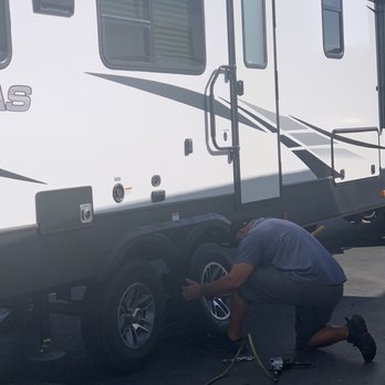 Beaver Tire - Thank you - A man is crouched down next to a white RV, using tools to change or repair the rear tire. The RV has multiple windows and a door, with a black lower section. The scene takes place outdoors on a paved street, with a yellow air hose and a tire-changing tool visible on the ground. - Tires Near Me - Beaver, Utah