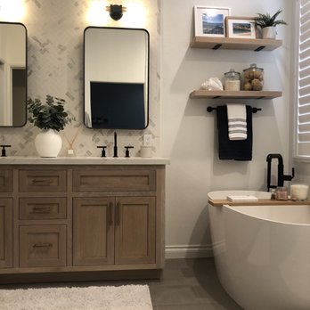 EDW Construction - Modern bathroom featuring a wooden vanity with black fixtures, two wall-mounted mirrors, and floating shelves with decor. A freestanding bathtub is positioned near a window with white shutters. - General Contractors Near Me