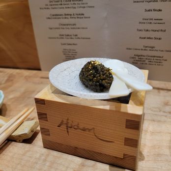 Hiden - Caviar - A small, textured white plate holds a serving of sea urchin roe (uni) accompanied by a dollop of white sauce. The plate is positioned on a wooden block with visible joinery, on a wooden table. In the background, a menu with various dishes and descriptions is partially visible, indicating a fine dining setting focused on Japanese cuisine. - Japanese Near Me - Miami, Florida