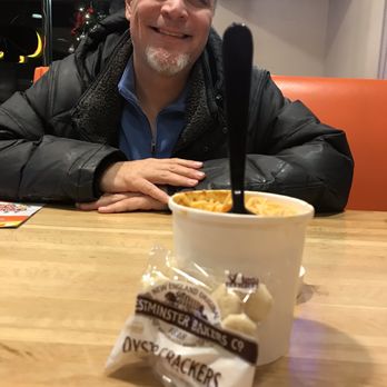 Meat & Potato Eatery-McHenry - Chili with cheese onions and oyster crackers - A man with gray hair and a beard sits at a table in a restaurant, smiling at the camera. In front of him is a cup of food with a black spoon and a packet of oyster crackers. - Sandwiches Near Me - Mchenry, Illinois