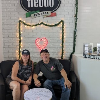 Freddo Midtown - a man and a woman sitting on a couch - Two individuals sit on a black sofa inside Freddo Midtown, with a white brick wall, festive decorations, and a sign reading 'Freddo' behind them. - Ice Cream & Frozen Yogurt Near Me - Miami, Florida