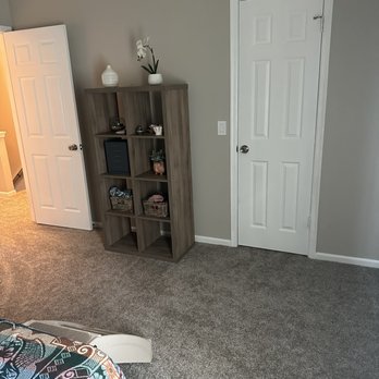J & B Carpets - The image shows a corner of a room with a gray wall, a closed white door, and a wooden bookshelf containing decorative objects and baskets. Part of a bed with a colorful quilt is visible in the lower left corner. The room has beige carpet flooring and a neutral color scheme. - Carpeting Near Me - Phoenix, Arizona