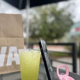 Mint and cucumber drink - A mint and cucumber beverage in a clear plastic cup is placed on a table outdoors, alongside a smartphone and a pink case.