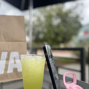 CAVA - Mint and cucumber drink - A mint and cucumber beverage in a clear plastic cup is placed on a table outdoors, alongside a smartphone and a pink case. - Salad Near Me - Spring, Texas