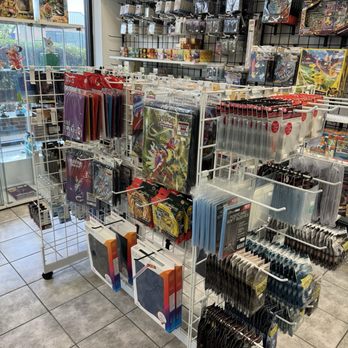 Curio Cavern - The image shows a retail store interior with a wire display rack holding various trading card game products, including booster packs, card sleeves, and accessories. The background includes shelves with boxed card sets, collectibles, and game-related merchandise. The store has large windows allowing natural light, and the floor is tiled. The display is organized with different products arranged for customer browsing, typical of a gaming or hobby store specializing in card games like Pokémon and Magic the Gathering. - Tabletop Games Near Me - Centreville, Virginia