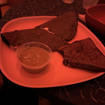 For All Things Good - I have no idea what this is. Maybe a cheese quesadilla - Three toasted bread slices with a small cup of green sauce on a white plate, set on a dark marble table in a dimly lit restaurant. - Cafes Near Me - Brooklyn, New York