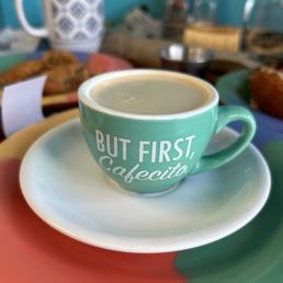 Cortadito - A ceramic coffee cup with the phrase "But First, Cafecito" printed on it, sitting on a saucer, with a blurred background of food and kitchen items.