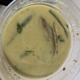 Green curry - The image shows a clear plastic container filled with green curry, featuring a creamy, yellowish-green sauce. Visible ingredients include pieces of vegetables and herbs, with some oil or liquid separation on the surface. The container is placed on a light-colored surface, and part of a black object is visible in the background.