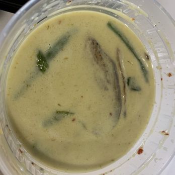 US Thai Cafe - Green curry - The image shows a clear plastic container filled with green curry, featuring a creamy, yellowish-green sauce. Visible ingredients include pieces of vegetables and herbs, with some oil or liquid separation on the surface. The container is placed on a light-colored surface, and part of a black object is visible in the background. - Thai Near Me - Edgewater, Colorado