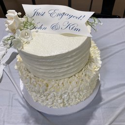 a wedding cake on a table - A two-tier wedding cake decorated with white frosting, floral accents, and a message topper that says "Just Engaged!" for John & Kim.