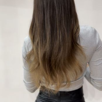 Salon Snob - A woman is shown from behind, with long, straight hair that transitions from dark brown at the roots to a lighter brown at the ends. She is wearing a light gray long-sleeve top and black leather pants. The background is plain and neutral, emphasizing her hair and outfit. - Hair Stylists Near Me - Chicago, Illinois