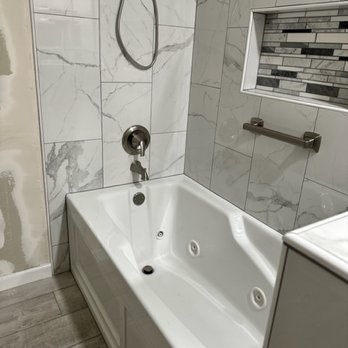 J J 'll Do It - The image shows a bathroom with a white, rectangular bathtub equipped with jets, set against a wall with large marble tiles. A small window with a mosaic tile border is visible above the bathtub, and a metal grab bar is mounted on the wall nearby. The floor appears to be tiled with wood-look tiles, and part of the bathroom wall on the left side shows unfinished or damaged drywall. - General Contractors Near Me - Clovis, California