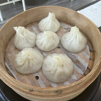 Super Bao - Xiao long bao (soup dumplings) - Six steamed soup dumplings (xiao long bao) are placed in a round bamboo steamer basket lined with parchment paper. The dumplings are white, pleated at the top, and appear freshly steamed. The basket is on a dark table, with a restaurant interior visible in the background, including chairs and a wooden floor. - Dim Sum Near Me - Mountain View, California