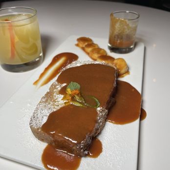 Sanctuary - Bread Pudding - A dessert plate displays a rectangular chocolate cake topped with caramel sauce and garnished with a small flower and mint leaf. The plate includes banana slices drizzled with caramel, a smear of caramel sauce, and powdered sugar. Two drinks are placed nearby: one with a yellowish liquid and a cherry, and another with a partially consumed beverage. The setting appears to be in a restaurant or dining area with a white table surface. - Cocktail Bars Near Me - Chicago, Illinois