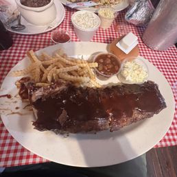 Ribs fries coleslaw and baked beans All decent. Fries tho were slightly better than the rest of the food - A large plate features barbecue ribs, French fries, baked beans, and coleslaw, with additional side dishes and condiments on a checkered tablecloth.