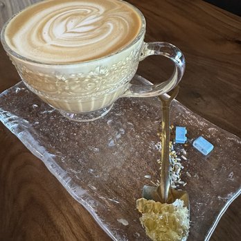 The Coffee Builders - Purple bee - A latte with foam art in a glass cup is placed on a textured glass plate on a wooden table. - Coffee & Tea Near Me - Phoenix, Arizona