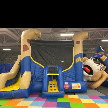 Bounce Mania - The image shows an inflatable bounce house with a slide and a clown figure at Bounce Mania in Tempe, AZ. The bounce house features a blue and yellow color scheme, with a black sliding surface and a small climbing area. The clown figure is part of the inflatable structure, positioned to the right. The setting includes colorful foam tiles on the floor and an indoor ceiling with visible lighting and ductwork. - Kids Activities Near Me - Tempe, Arizona