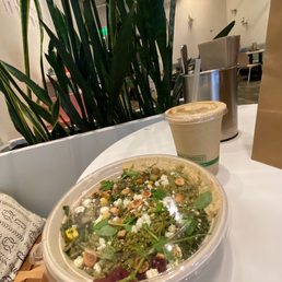New* Pesto Power Bowl - A salad with mixed greens, nuts, cheese, and other toppings is in a clear plastic container on a white table. Next to it is a smoothie in a plastic cup with a straw. The background shows indoor seating with chairs, tables, and decorative plants, indicating a modern restaurant environment.