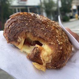 Ham and Cheese Croissant - A croissant with a crispy, layered exterior and sesame seeds on top, filled with melted cheese and ham, partially eaten. The croissant is held on a white napkin, with an outdoor urban background featuring buildings and trees.