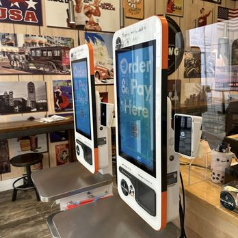 Sweeting - Kiosk - Two self-service ordering kiosks with large screens displaying 'Order & Pay Here' are situated on a stainless steel counter inside a restaurant. The kiosks have card readers and touchscreens for customer use. Behind the kiosks, a wall is decorated with vintage posters, artwork, and photographs, creating a retro ambiance. The setting includes a wooden countertop with a small container holding utensils and a clear divider, indicating a dine-in or takeout environment. - Bubble Tea Near Me - Ann Arbor, Michigan