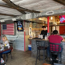 people sitting at the bar - A casual brewery interior featuring a bar area with patrons seated, a chalkboard menu, and a large American flag on the wall.
