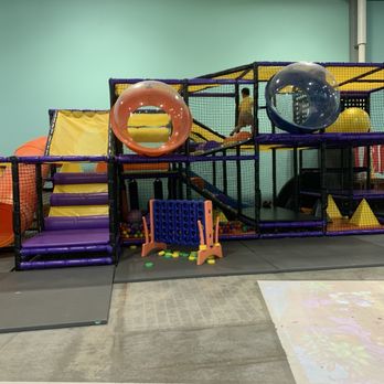 Wisco Kidz Place - The image shows an indoor children's play area with a multi-level play structure enclosed by netting. It includes slides, tunnels, and ball pits, with a soft, padded floor for safety. A small blue and orange connectable game is placed in front of the structure, and a child is visible playing inside the structure. The setting appears to be in a recreational or entertainment facility designed for kids. - Playgrounds Near Me - Madison, Wisconsin
