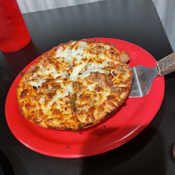 Pizza 9 - All meat individual pizza - A pizza topped with melted cheese and meat, possibly sausage and pepperoni, is placed on a red plastic plate. The pizza has a golden-brown crust and is partially sliced, with a metal pizza cutter resting on the plate. The setting appears to be a casual dining area with a black table and a corrugated metal wall in the background. - Pizza Near Me - Grants, New Mexico