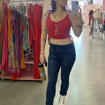 Indy clover - A woman in a clothing store poses for a mirror selfie, dressed in a red crop top, dark jeans, and white boots, with sunglasses hanging from her top. - Thrift Stores Near Me - Plano, Texas