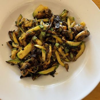 Tune-Up Cafe - Sautéed vegetables - A white ceramic bowl holds a serving of sautéed vegetables, including sliced zucchini, mushrooms, and other mixed vegetables. The dish appears to be cooked with some seasoning and oil, and is placed on a wooden table surface. The vegetables are cut into strips and are slightly browned from cooking. - New Mexican Cuisine Near Me - Santa Fe, New Mexico