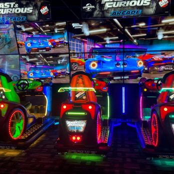 Dave & Buster's Schaumburg - The image shows a racing arcade game setup with three brightly colored racing seats equipped with steering wheels and controls. Each seat has illuminated accents in green, red, and purple, and displays the Fast & Furious arcade logo. Behind the seats, multiple screens display gameplay scenes from the Fast & Furious arcade game, featuring cars and racing environments. The setup is located indoors, likely in an arcade or entertainment center, with vibrant lighting and a themed racing environment. - Sports Bars Near Me - Schaumburg, Illinois