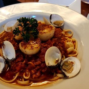 MIX Kitchen & Bar - Clams with red sauce and added 3 scallops ... DELICIOUS! - A white plate holds a serving of pasta with a red clam sauce, garnished with several seared scallops and a sprig of parsley. Clams in shells are arranged around the scallops, and the dish is set on a wooden table with a glass of dark beverage and an empty plate nearby. - Cocktail Bars Near Me - Ithaca, New York
