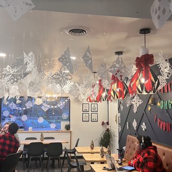 Better Together Cafe - Elf themed Christmas decorations - The cafe interior features white paper snowflakes hanging from the ceiling and red bows on light fixtures, creating a festive Christmas atmosphere. There are several tables with small Christmas tree decorations, and a woman wearing headphones and a red plaid shirt is seated at one table working on a laptop. The decor includes a window with holiday-themed decorations and framed pictures on the wall. The ceiling has an industrial duct, and the overall setting is cozy and seasonal. - Desserts Near Me - South Milwaukee, Wisconsin