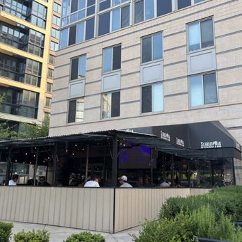 Scarlet Oak - Outdoor seating at Scarlet Oak restaurant and bar in Washington, DC, features a covered patio with multiple patrons seated at tables. The patio is enclosed by a low wall, and the restaurant's black awning displays its name. The background includes a multi-story building with large windows, and surrounding greenery is visible in the foreground. - New American Near Me - Washington, DC, District of Columbia