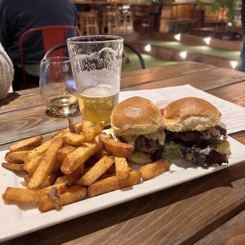 The Jack - Beef Sliders - A rectangular white plate holds two sliders with buns, beef patties, and lettuce, accompanied by seasoned fries. A partially filled glass of beer and a small glass of white wine are placed behind the plate. The background shows a rustic restaurant interior with wooden furniture, warm lighting, and patrons seated at tables. - American Near Me - Camden, Maine