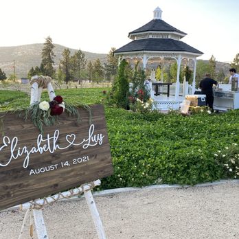 Willow Tree Weddings - A wooden wedding sign with white cursive lettering shows the names Elizabeth and Luis, along with the wedding date August 14, 2021. The sign is decorated with flowers and greenery at the top and is mounted on a white stand. The setting is an outdoor garden with lush green plants, a white gazebo, and a group of people near a food or drink station, with mountains and trees in the background. - Wedding Planning Near Me