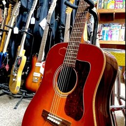 New to me Guild. - A sunburst acoustic guitar is positioned in a music store, surrounded by electric guitars, guitar cases, and musical accessories on shelves and hooks.