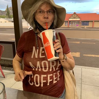 The Human Bean - Diana enjoying her Frozen blended Salted Carmel,Raspberry & Almond, my recipe - A woman with gray hair, glasses, and a wide-brimmed hat is holding a large Coca-Cola cup with a straw, sipping her drink. She wears a maroon T-shirt with the words 'NE MORE COFFEE' printed on it and has a beige bag over her shoulder. The background shows a window with an outdoor view of a street and a building with a red roof, under a cloudy sky. - Coffee & Tea Near Me - Phoenix, Arizona