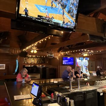 John B's - Bar - A bar interior with a large TV displaying a basketball game, patrons seated at the bar, some using phones, and arcade machines in the background. - Dive Bars Near Me - Pantego, Texas