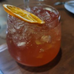 A glass of iced cocktail with an orange slice garnish is placed on a wooden table, with a blurred background.
