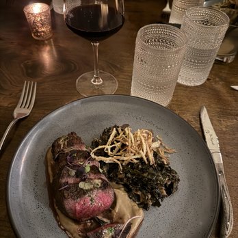 Eberly - Waygu steak strip - A dinner setting with a plate of sliced steak, cooked greens topped with crispy onions, a glass of red wine, and three glasses of water on a wooden table. - Bars Near Me - Austin, Texas