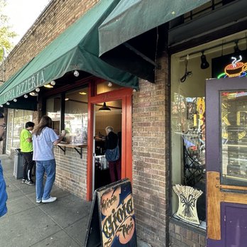 Gioia Pizzeria - Outside - The image shows several people standing outside Gioia Pizzeria in Berkeley, CA, on a sidewalk. The pizzeria has a green awning with the name partially visible, and a red door leading inside. A signboard on the sidewalk advertises slices of pizza. The storefront features brick walls, and the interior is visible through the window, with a person inside. The scene is set during daytime with trees visible in the background. - Pizza Near Me - Berkeley, California