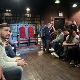 People are seated in a theater setting with a throne and a large bookshelf backdrop, preparing for a performance.
