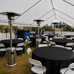 Elegant outdoor tent setup with round tables, black tablecloths, heaters, and white folding chairs.