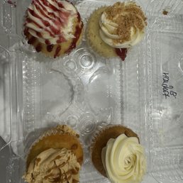 Red Velvet French Toast - Four cupcakes are placed inside a clear plastic container, each in a paper liner. The cupcakes have different toppings: one with white frosting and red syrup drizzle, another with white frosting and brown crumbs, a third with caramel-colored frosting and caramel drizzle, and the last with white frosting piped in a swirl. The container is on a white surface.