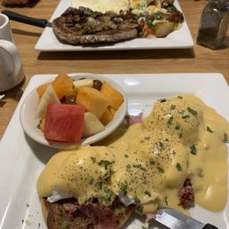 Eggs bennie and a pork steak and eggs - Eggs Benedict topped with hollandaise sauce and garnished with herbs, served on a plate with a side of mixed fresh fruit.