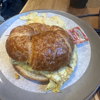 Black Lion Cafe - A breakfast sandwich on a tray includes scrambled eggs, melted cheese, and a flaky croissant bun. Accompanying the sandwich are ridged potato chips and a small packet of Smucker's strawberry jam. The tray is placed on a wooden table, with a black cup and a backpack visible in the background. - Cafes Near Me - Silver Spring, Maryland