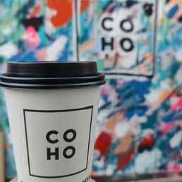 White Hot Chocolate - A person holds a paper coffee cup featuring the COHO logo and the phrase 'Socially Good Coffee' in front of a colorful, abstract mural with splashes of blue, red, orange, and pink. The mural includes the COHO logo within a square outline, matching the logo on the cup.