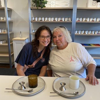 AURA Candle Bar - Mom and I had a blast here!!! - Two women sit at a table in a candle studio, with candles, tasting spoons, and trays in front of them, surrounded by shelves of candles. - Candle Stores Near Me - Chicago, Illinois