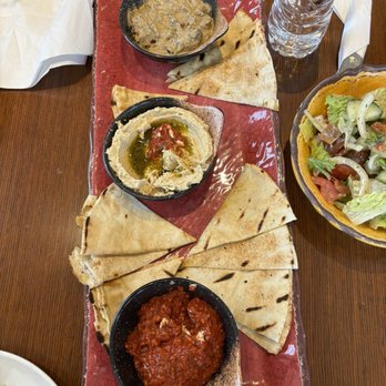 Charr'd - Trio Dip Plate - A rectangular tray holds three bowls of Indian appetizers: hummus with olive oil, a chutney, and a vegetable dip, accompanied by naan bread. A side salad and bottled water are also visible. - Mediterranean Near Me - Toronto, Ontario