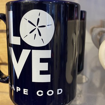 Cool as a Moose - The image shows a black ceramic mug with a white graphic design. The design includes the word "LOVE" with a snowflake symbol replacing the letter "O". Below the word, the phrase "SHAPE COD" is printed in smaller white letters. The mug is placed on a surface with other objects visible in the background, indicating it is in a retail or display setting. - Women's Clothing Near Me - Provincetown, Massachusetts
